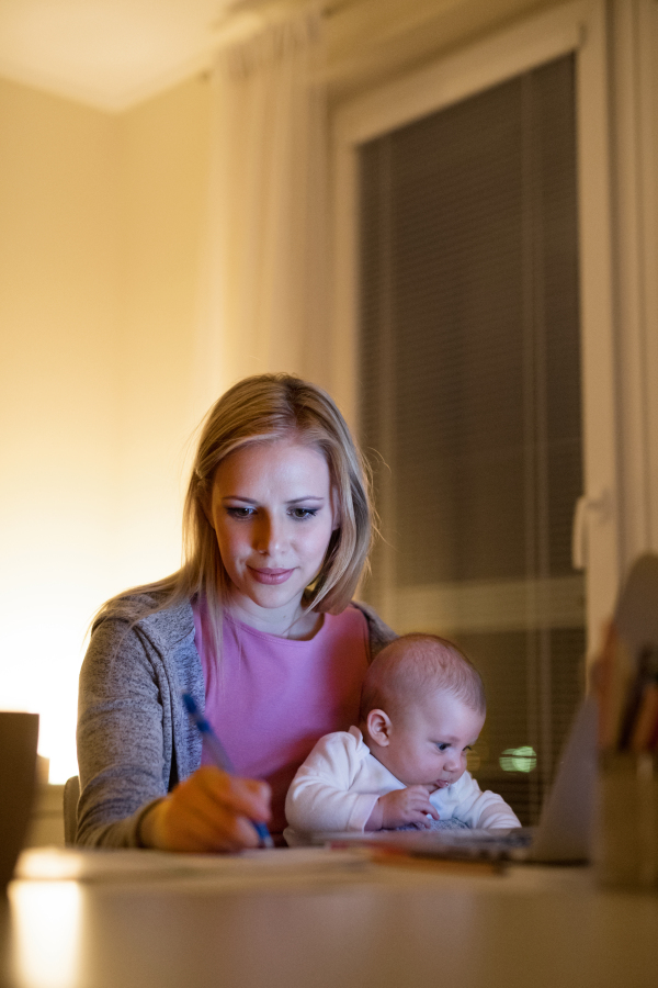 Beautiful young mother at home at night with her little baby son in the arms, working on laptop