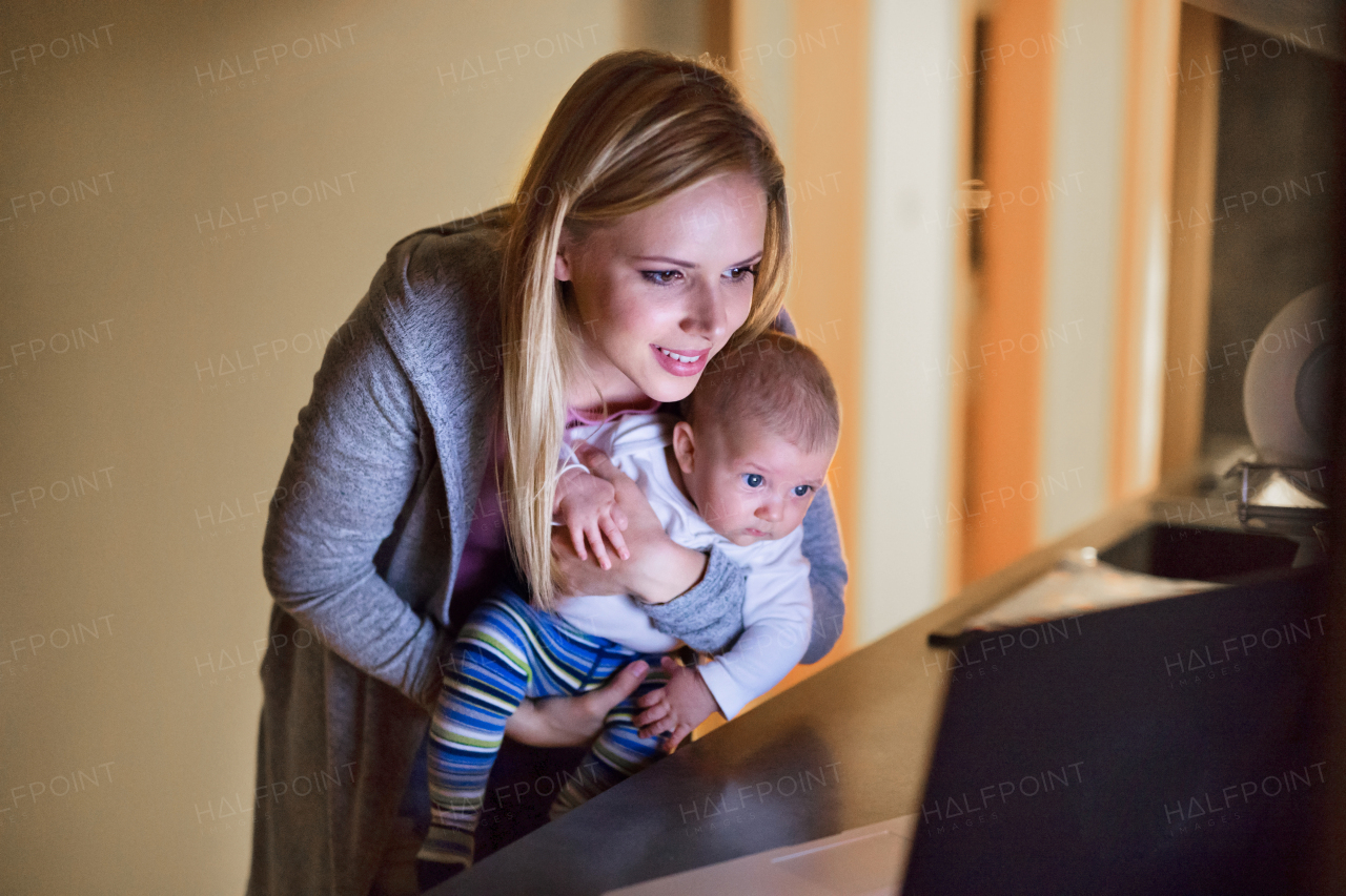 Beautiful young mother at home at night with her little baby son in the arms, laptop in front of her