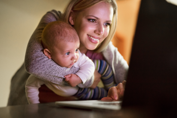 Beautiful young mother at home at night with her little baby son in the arms, working on laptop