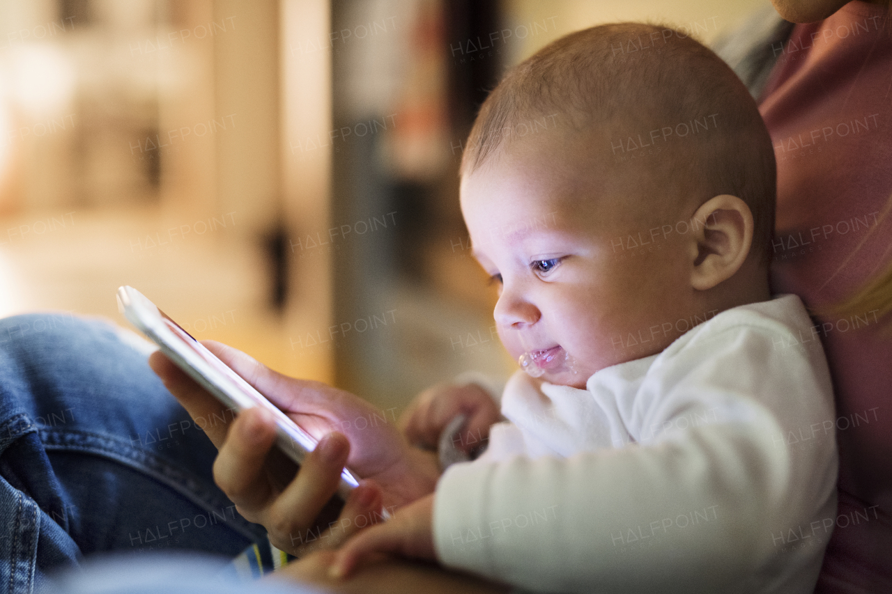 Unrecognizable young mother at home with her little baby son in the arms, sitting on the kitchen floor, holding smart phone