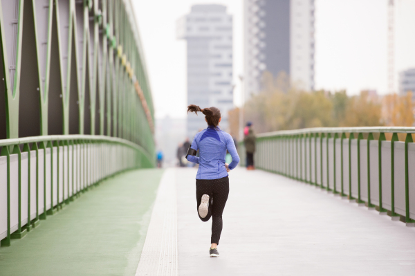 Beautiful young woman with smart phone in the city, tracking her progress, running on green steel bridge. Rear view.