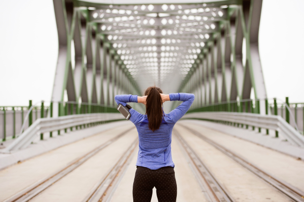 Beautiful young runner with smart phone in the city, tracking her progress, warming up and stretching on green steel bridge. Rear view.