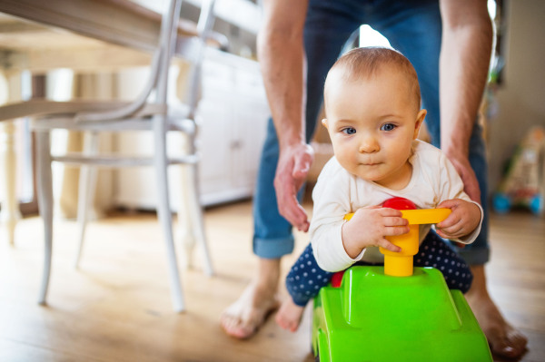 Unrecognizable father with a baby girl sitting on a toy car at home. Paternity leave.