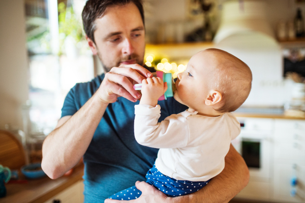 A father with a baby daughter at home. A cute girl drinking water from the bottle. Paternity leave.