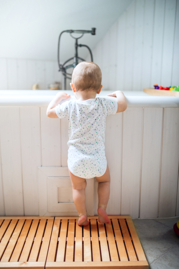 A toddler child standing at the bath in the bathroom at home. Rear view.