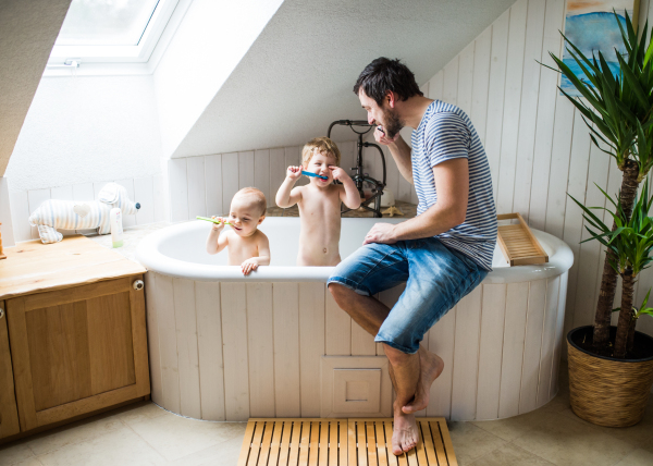 Father with two toddlers in the bath tub brushing teeth in the bathroom at home. Paternity leave.