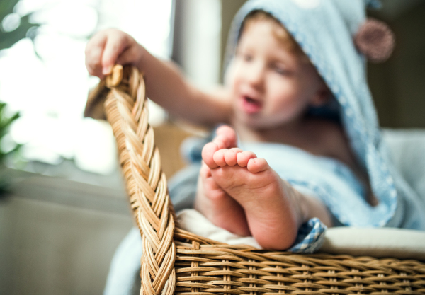 A toddler boy wrapped in a towel sitting on the wicker chair in the bathroom at home.