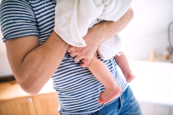 Unrecognizable father with a toddler child wrapped in a towel in a bathroom at home. Paternity leave.