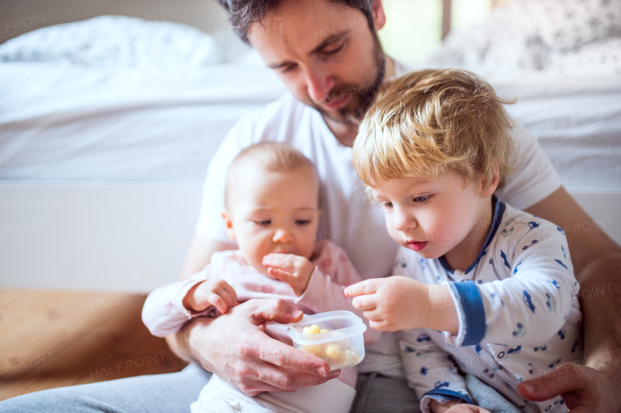 Handsome father with toddler children eating finger food in bedroom at home. Paternity leave.