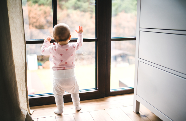 A toddler child standing by the window on the wooden floor in bedroom at home. Rear view.