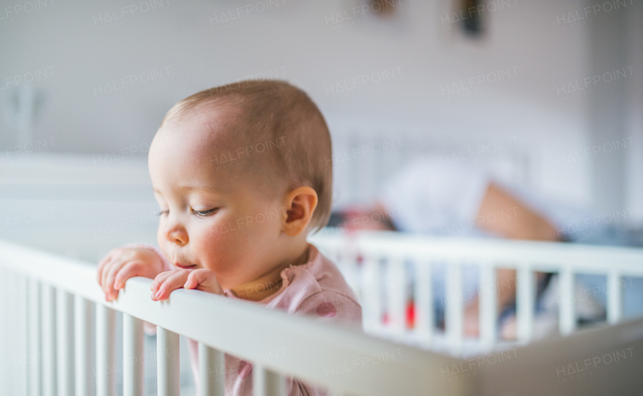 A happy toddler girl standing in a cot in the bedroom at home. Unrecognizable father in the background. Close up.