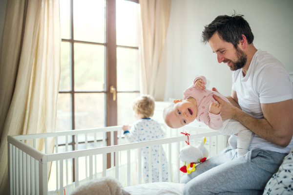 A mature father with toddler children in bedroom at home at bedtime. Paternity leave. A man holding little girl, a boy standing in a cot.