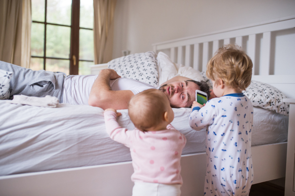 Two toddler children playing with smartphone and their father lying on bed in the bedroom. Paternity leave.