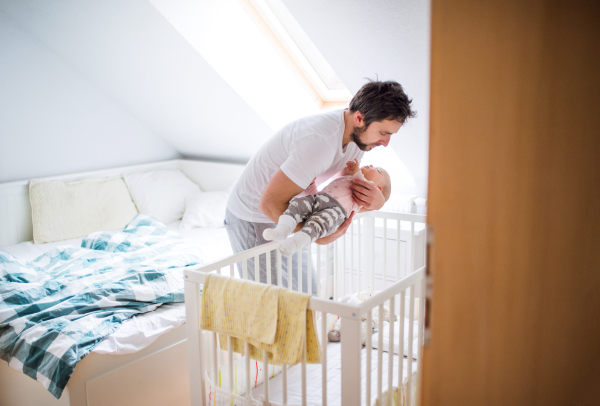 Mature father putting a sleeping toddler girl into a cot at home. Paternity leave.