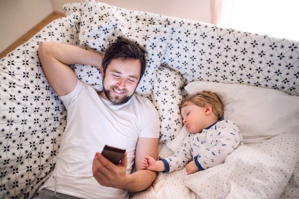 Father with smartphone and a sleeping toddler boy in bed at home. Paternity leave.