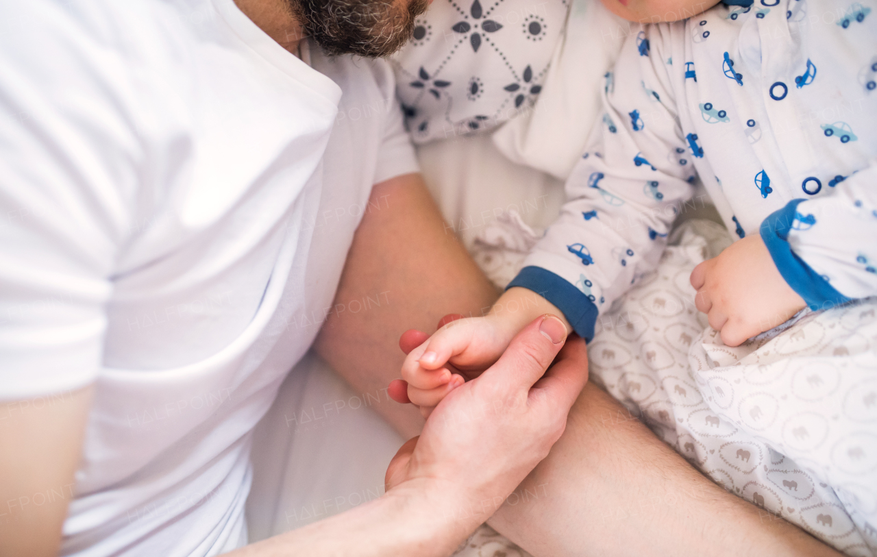 Unrecognizable father holding hand of a sleeping toddler boy lying in bed at home. Paternity leave.