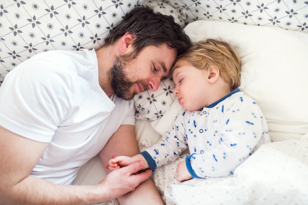 Father holding hand of a sleeping toddler boy lying in bed at home. Paternity leave.