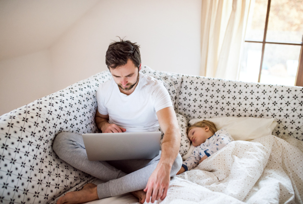 Father with laptop and a sleeping toddler boy in bed at home. Paternity leave.