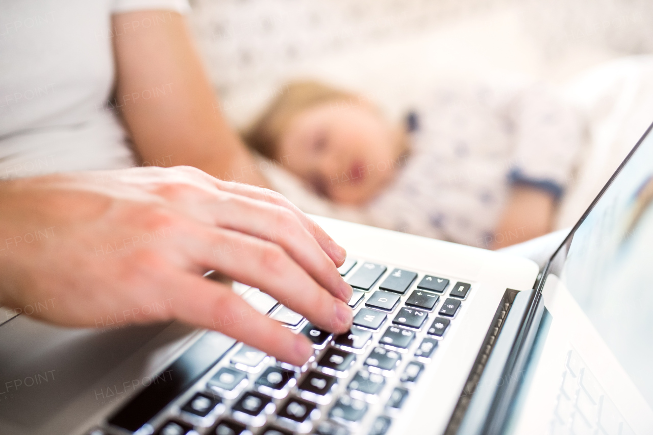 Unrecognizable father with laptop and a sleeping toddler boy in bed at home. Paternity leave.