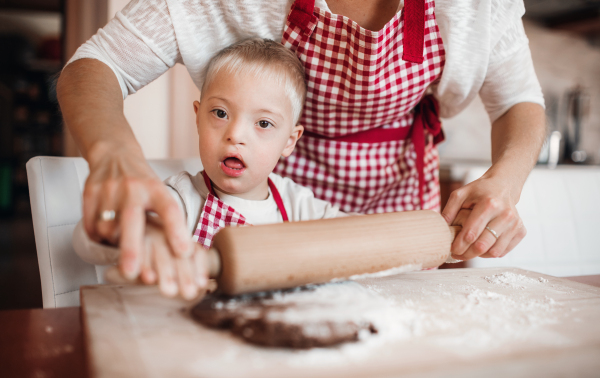 A handicapped down syndrome child and his unrecognizable mother with checked aprons indoors baking in a kitchen.