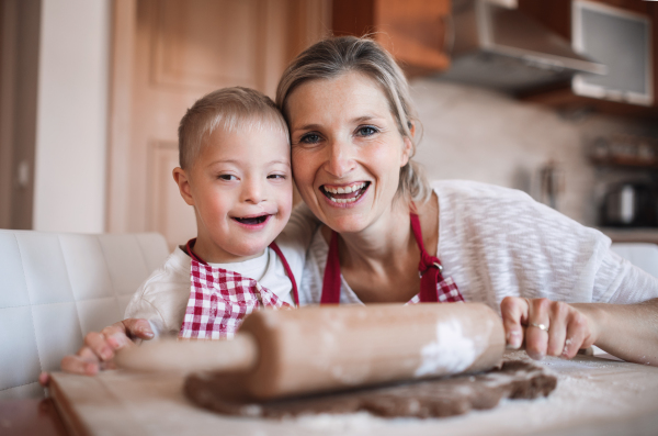 A portrait of handicapped down syndrome boy and his mother with checked aprons indoors baking in a kitchen.