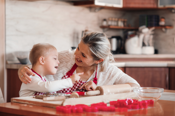 A happy handicapped down syndrome child and his mother with checked aprons indoors baking in a kitchen, having fun.