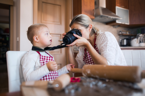 A handicapped down syndrome boy and his mother indoors in a kitchen taking photos with a digital camera when baking.