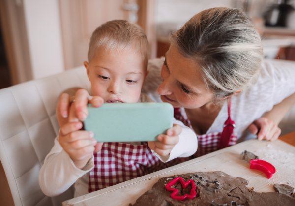 A handicapped down syndrome child and his mother with smartphone indoors in a kitchen, taking selfie when baking.