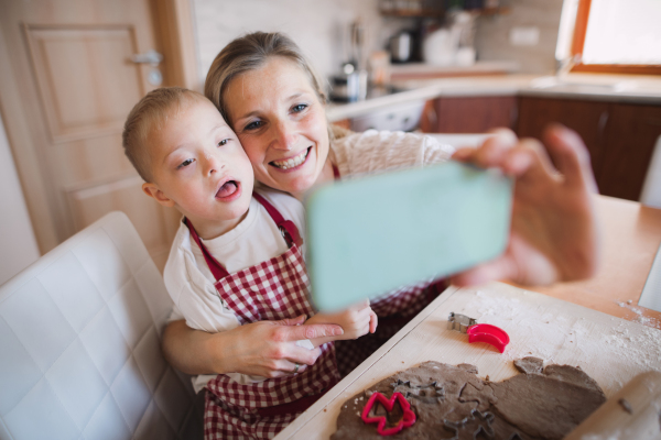 A handicapped down syndrome boy and his mother with smartphone indoors in a kitchen, taking selfie when baking.