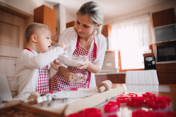 A handicapped down syndrome child and his mother with checked aprons indoors baking in a kitchen.