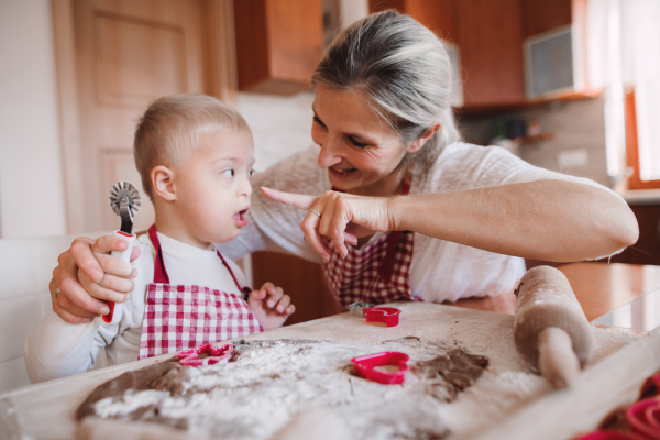 A happy handicapped down syndrome child and his mother with checked aprons indoors baking in a kitchen, having fun.