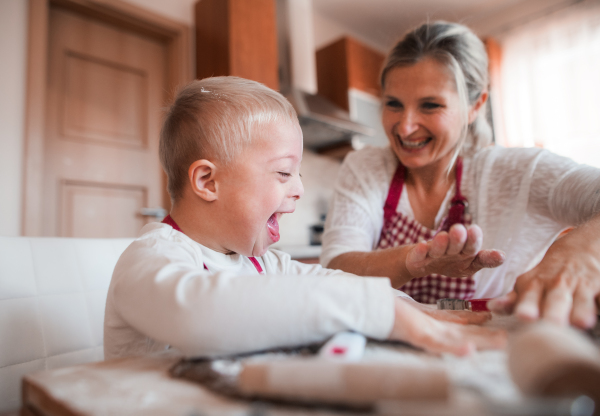 A laughing handicapped down syndrome child and his mother with checked aprons indoors baking in a kitchen, having fun.