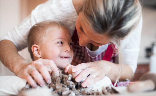 A happy handicapped down syndrome child and his mother indoors baking in a kitchen. A close-up.