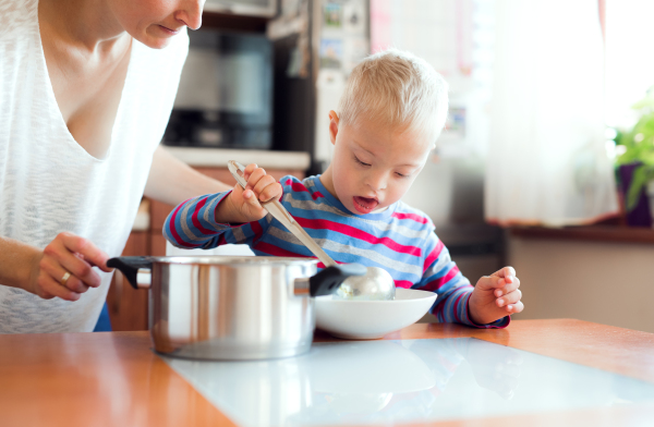 A happy handicapped down syndrome boy with unrecognizable mother pouring soup in a plate indoors, lunch time.