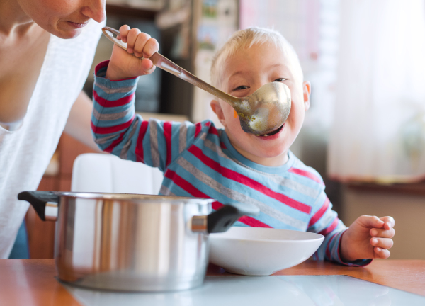 A happy handicapped down syndrome boy eating soup from a ladle indoors, lunch time.