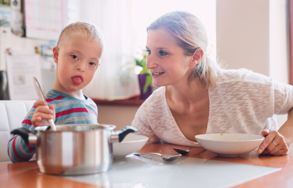 A handicapped down syndrome child with his mother indoors eating lunch or dinner.