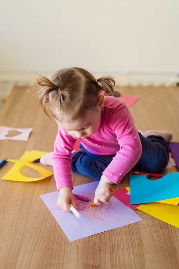 Cute little girl sitting on a floor at home drawing on colorful papers