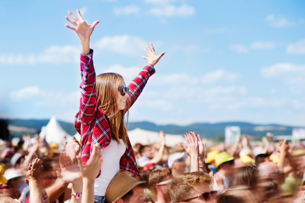 Teenage couple at summer music festival under the stage in a crowd enjoying themselves. Handsome man giving beautiful young woman piggyback, arms in the air.