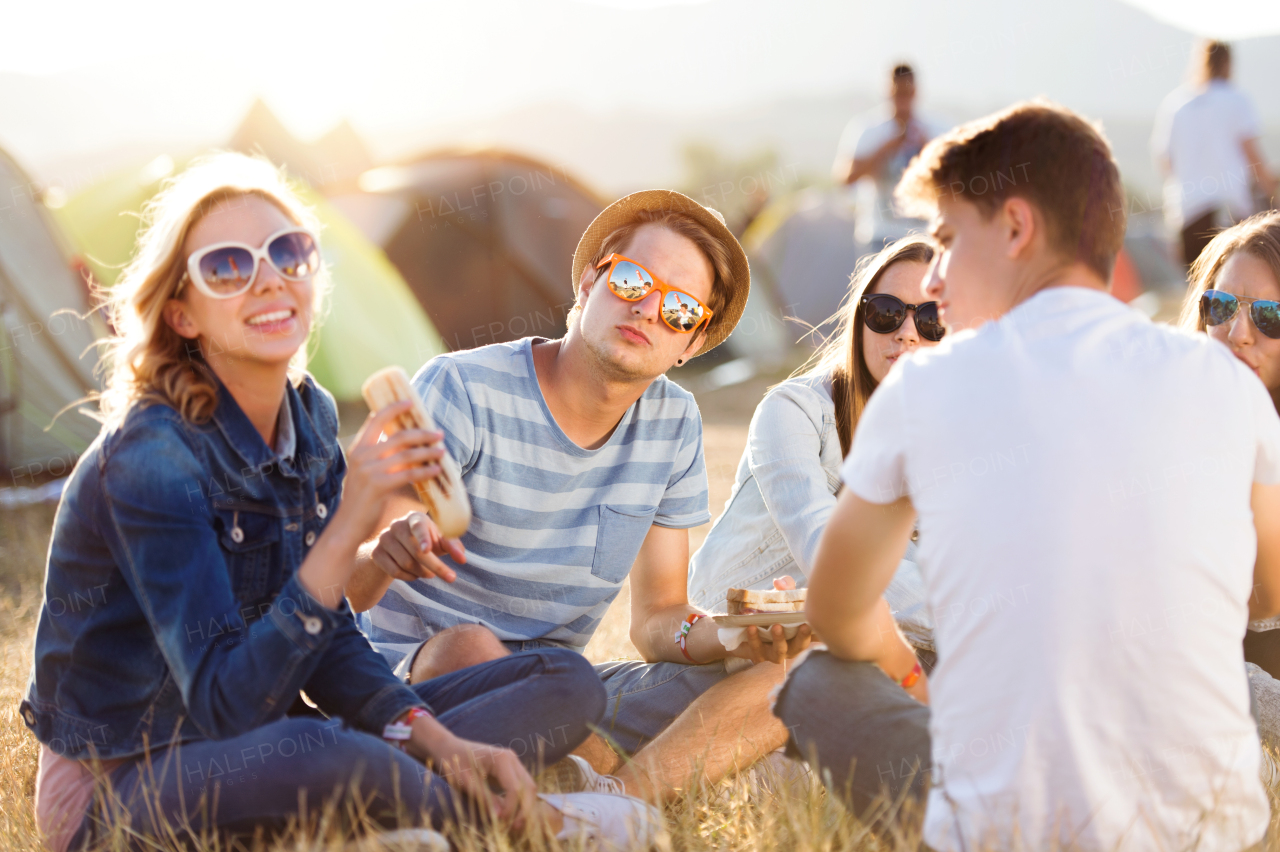 Group of teenage boys and girls at summer music festival, sitting on the ground in front of tents resting and eating