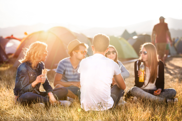 Group of teenage boys and girls at summer music festival, sitting on the ground in front of tents resting and eating