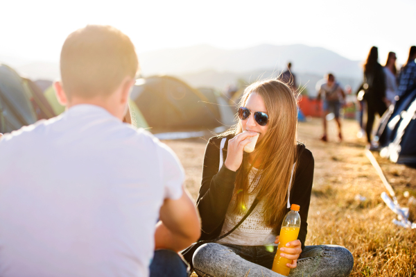 Group of teenage boys and girls at summer music festival, sitting on the ground in front of tents eating and drinking