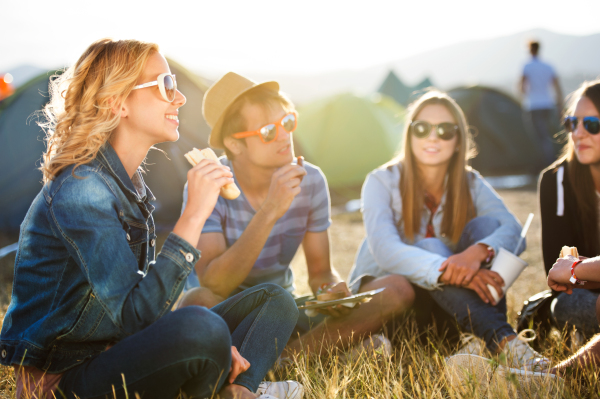 Group of teenage boys and girls at summer music festival, sitting on the ground in front of tents eating