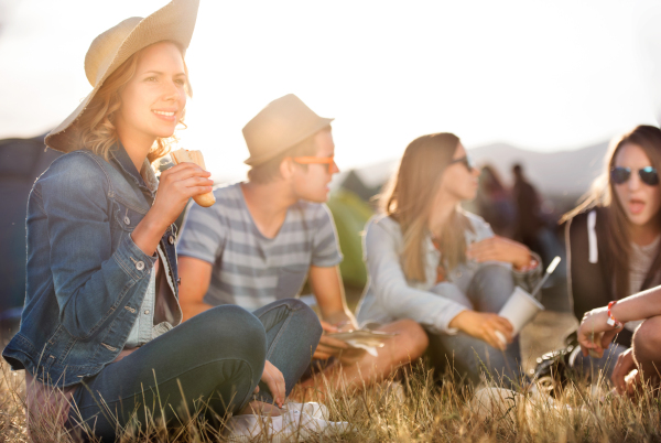 Group of teenage boys and girls at summer music festival, sitting on the ground in front of tents resting and eating