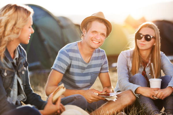 Group of teenage boys and girls at summer music festival, sitting on the ground in front of tents, resting and eating