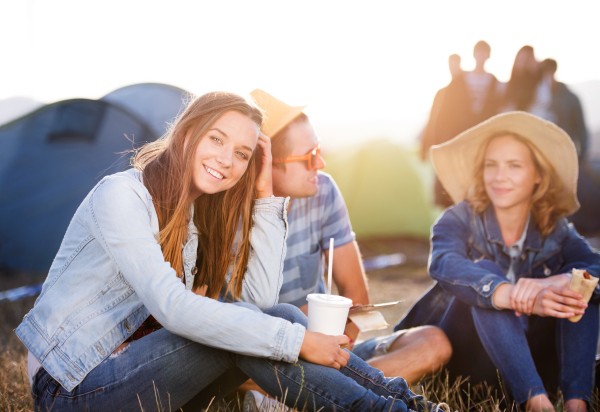 Group of teenage boys and girls at summer music festival, sitting on the ground in front of tents, resting and eating