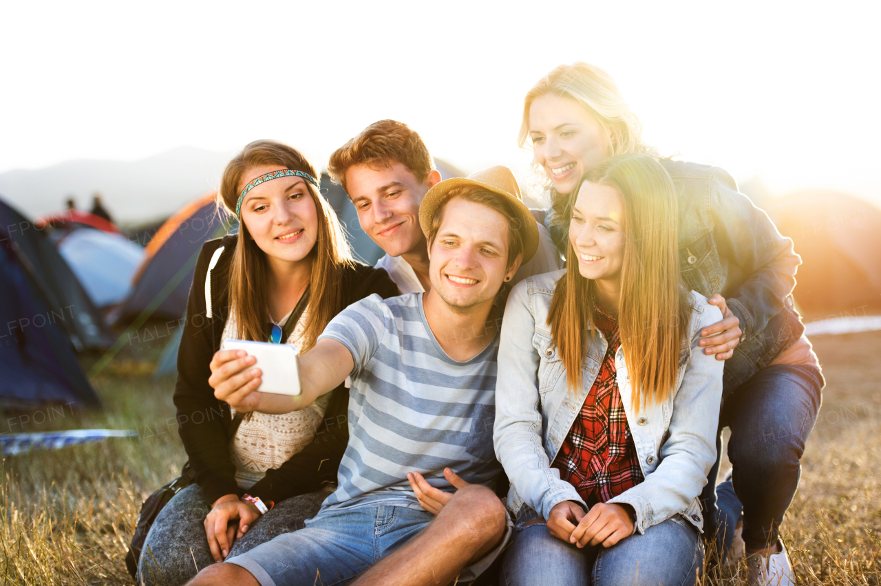 Group of teenage boys and girls at summer music festival, sitting on the ground, taking selfie with smart phone