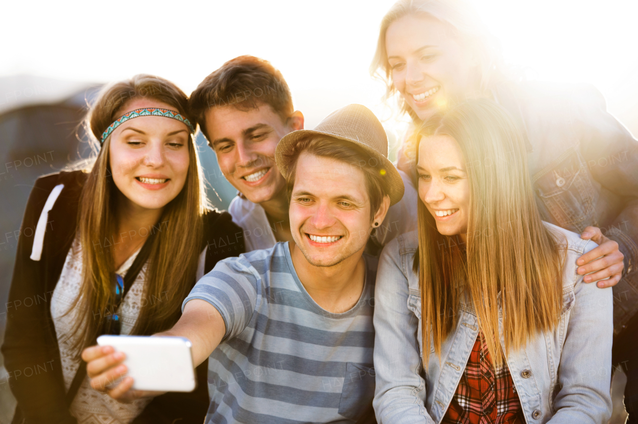 Group of teenage boys and girls at summer music festival, sitting on the ground, taking selfie with smart phone