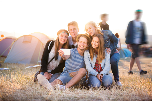 Group of teenage boys and girls at summer music festival, sitting on the ground, taking selfie with smart phone