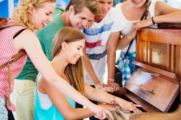 Group of teenage boys and girls at summer music festival, beautiful young woman plays the piano. Sunny day.