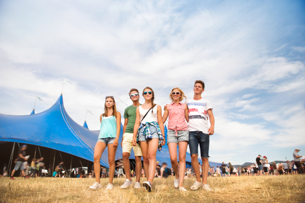 Group of teenage boys and girls at summer music festival walking in front of big blue tent, sunny day